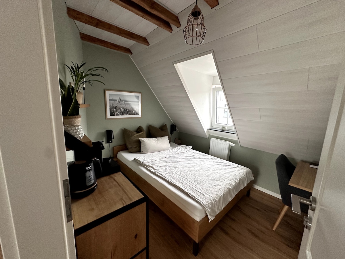 The guest room features a spacious bed with a light-colored bedding set, accompanied by decorative pillows. A simple wooden desk and chair are positioned beside a window, while a plant adds a touch of greenery. Exposed beams enhance the room's character.