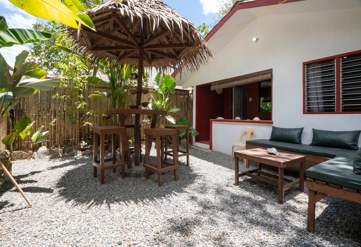 A shaded native high table and chairs provide a gathering space in the fenced garden, surrounded by lush greenery. Nearby, a comfortable seating area with cushions overlooks the gravel ground. The bungalow exterior features bright red accents and large windows, inviting natural light.