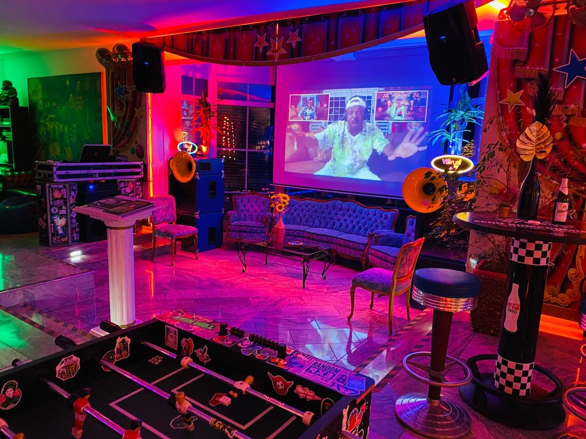A vibrant entertainment area is highlighted by colorful lighting and a large screen displaying content. A foosball table can be seen in the foreground, while retro-style sofa seating and a bar area create a welcoming atmosphere for social gatherings.