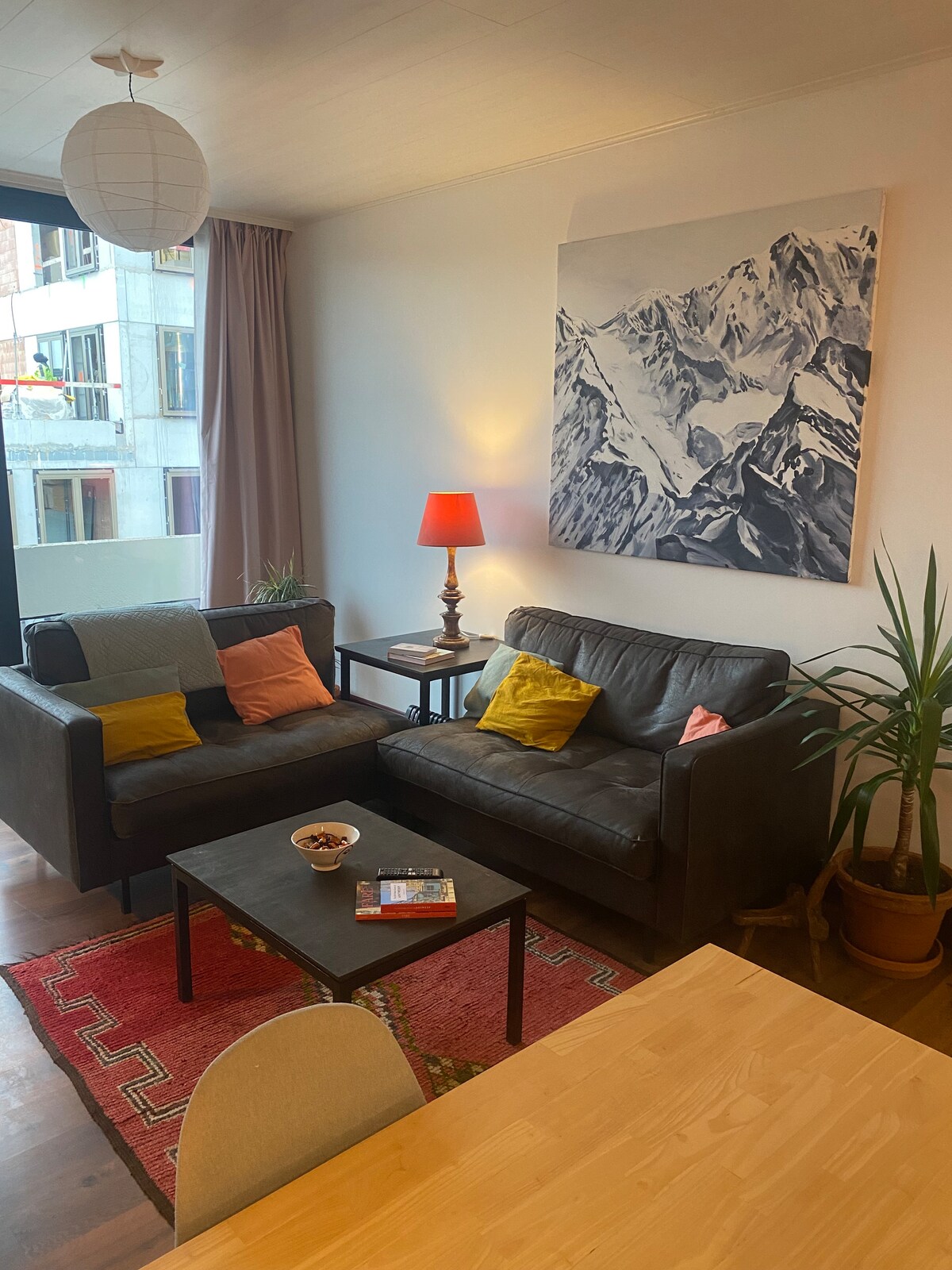 The living area features a dark leather sofa adorned with colorful cushions, accompanied by a small coffee table. A bright lamp casts a warm glow, complementing a mountain-themed artwork on the wall. A plant adds a touch of greenery, enhancing the bright atmosphere.