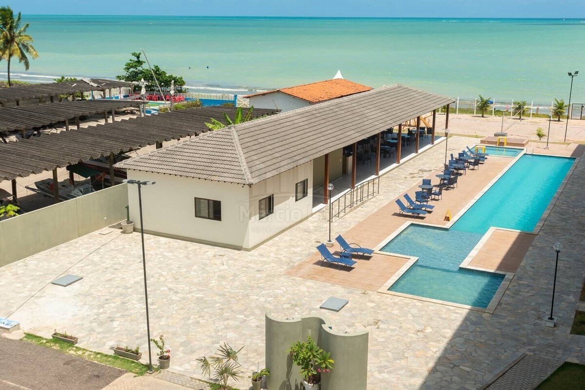 The outdoor area showcases a long swimming pool lined with lounge chairs. A spacious building is visible alongside the pool, with a covered patio offering shade. The ocean can be seen in the background, enhancing the seaside environment.
