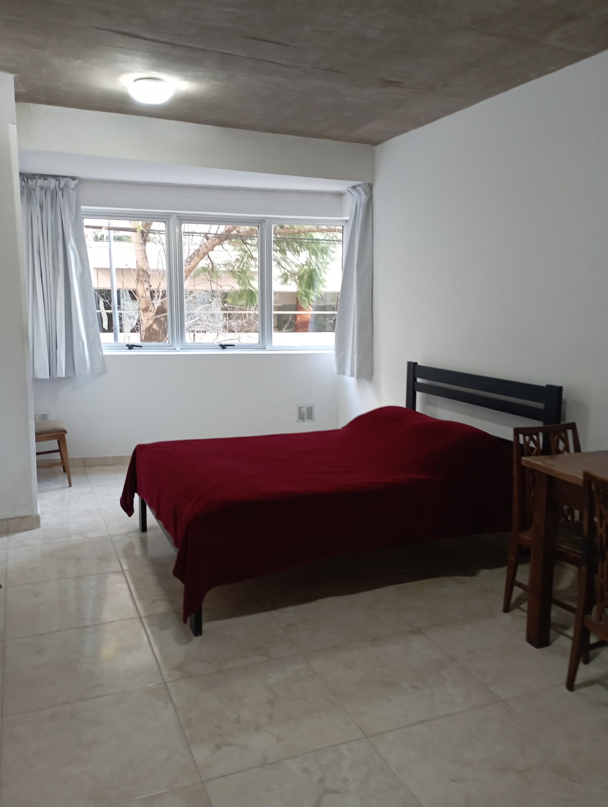 A simple room is presented with a spacious bed draped in a deep red cover. Natural light enters through two large windows, framed by light curtains. A small dining area features a wooden table and four chairs, set against a neutral wall.