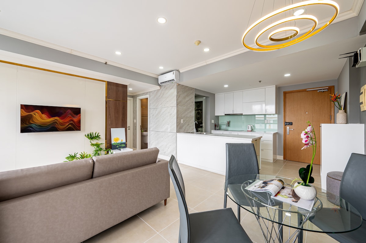 A modern living area features a comfortable gray sofa, positioned towards a sleek white kitchen with glossy cabinets. A dining table with elegant chairs is visible nearby. Decorative elements include a wall-mounted television and a plant, contributing to a stylish yet functional atmosphere.