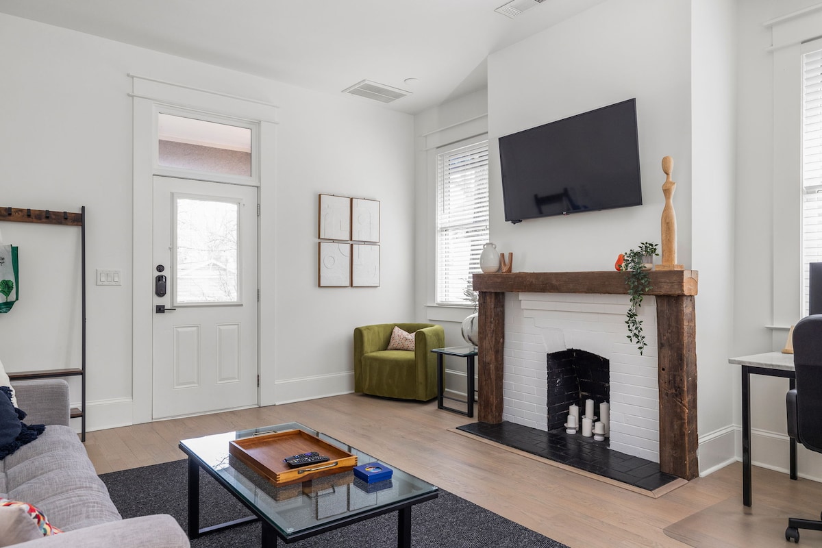 The living room features a modern aesthetic with a gray sofa, green armchair, and a glass coffee table. A rustic wooden fireplace serves as a focal point, complemented by a flat-screen television mounted above. Natural light fills the space through tall windows.