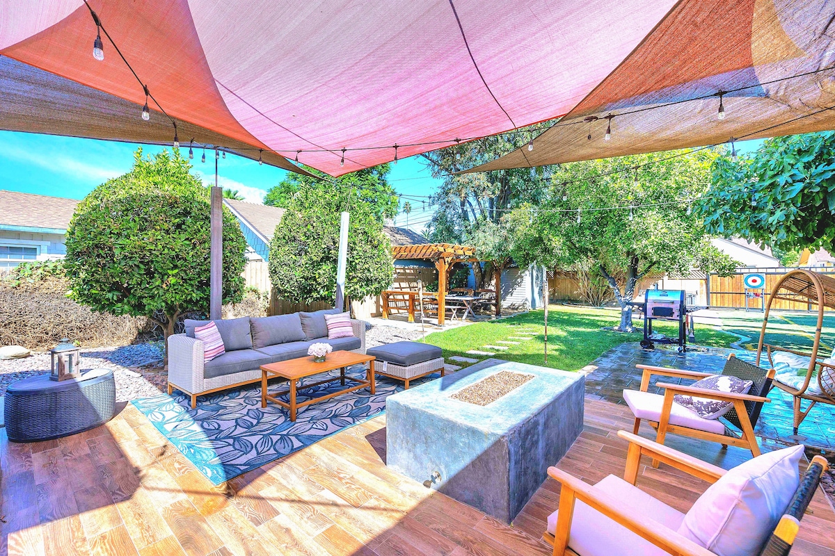 An outdoor seating area is shaded by large canopies, featuring a comfortable sofa and wooden coffee table. A firepit is positioned on a wooden deck, surrounded by lounge chairs. Lush greenery and decorative elements are visible in the background, creating a relaxed outdoor environment.