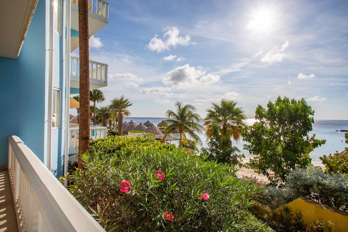Bright sunlight illuminates a balcony view overlooking lush greenery and vibrant flowers. The beach and ocean are visible in the background, framed by palm trees and shaded cabanas. A clear sky complements the serene setting, creating an inviting atmosphere for relaxation.