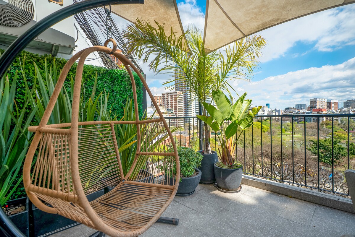 A comfortable hanging chair is featured on a spacious terrace surrounded by lush greenery. Planters with vibrant plants and trees enhance the outdoor space. A view of the city skyline and nearby park is visible in the background, creating a tranquil outdoor retreat.