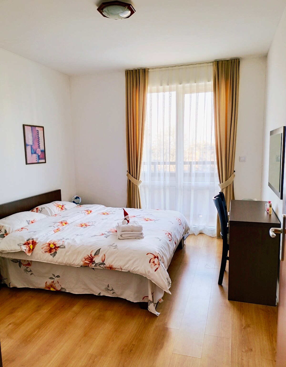 A well-lit bedroom features a comfortable bed with floral bedding and a neatly arranged towel. A desk and chair are positioned against the wall, while natural light streams through a large window draped with sheer curtains.