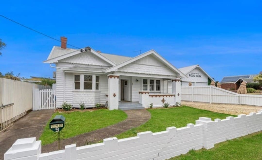 The exterior of a charming white house with a classic design is showcased, surrounded by a well-maintained lawn. A low white fence encloses the property, and a well-defined pathway leads to the entrance. Bright blue skies enhance the inviting atmosphere.