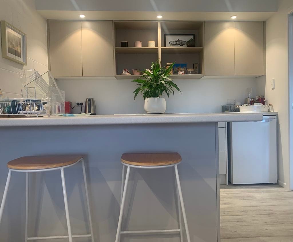 A modern kitchenette is equipped with essential appliances, including a kettle and a refrigerator. The light-colored cabinetry is complemented by overhead shelves that display decorative items. Two minimalist stools are positioned at the counter, where soft lighting creates a welcoming atmosphere.