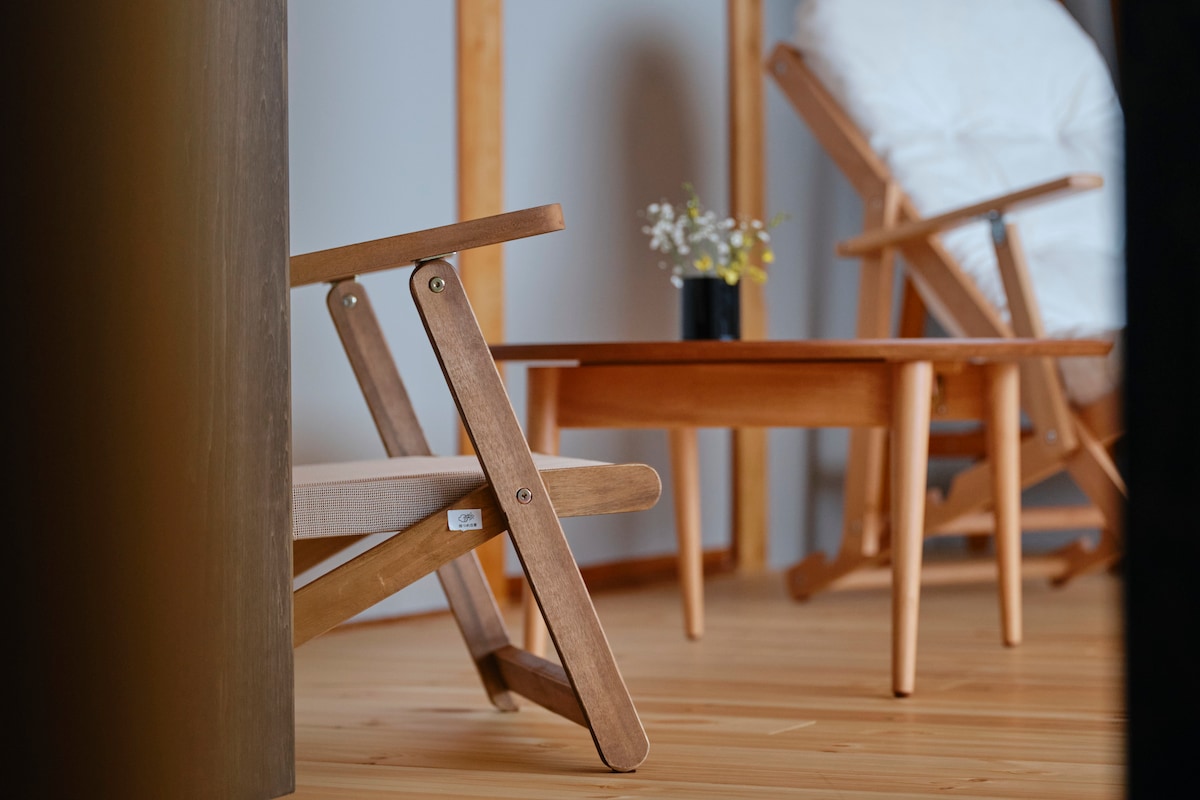 A cozy seating area is showcased, featuring a wooden folding chair and a comfortable armchair. A small wooden table is situated beside them, adorned with a vase holding fresh flowers. The warm wooden flooring adds to the inviting simplicity of the space.