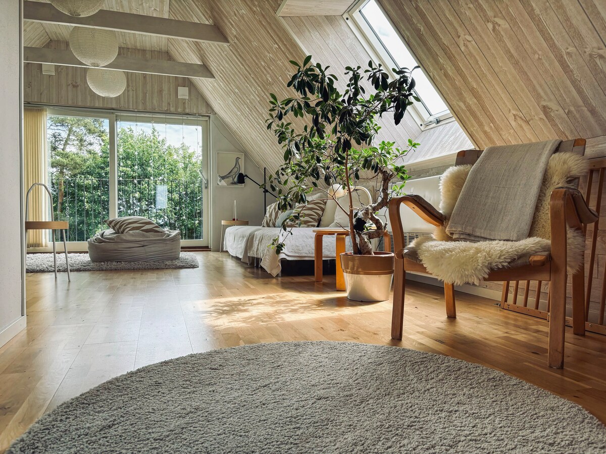 An inviting upper living area is illuminated by natural light from large, angled windows. A comfortable seating area features a wooden chair with a light blanket, alongside a soft bean bag. Lush greenery from potted plants adds a touch of nature, complemented by a round area rug.