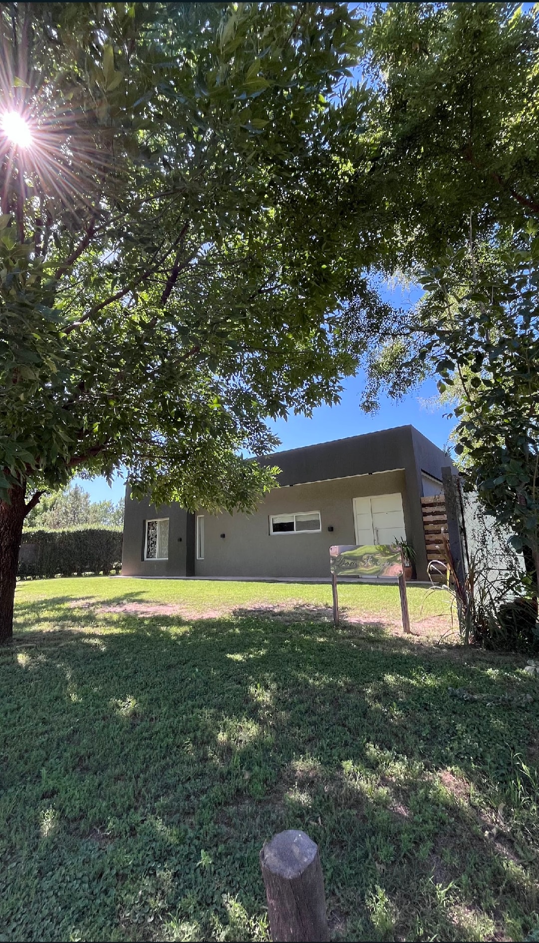 A modern single-story home is surrounded by lush greenery. Sunlight filters through the leaves of nearby trees, casting gentle shadows on the grassy area. A welcoming entrance door is visible, alongside a sign indicating the property's name.