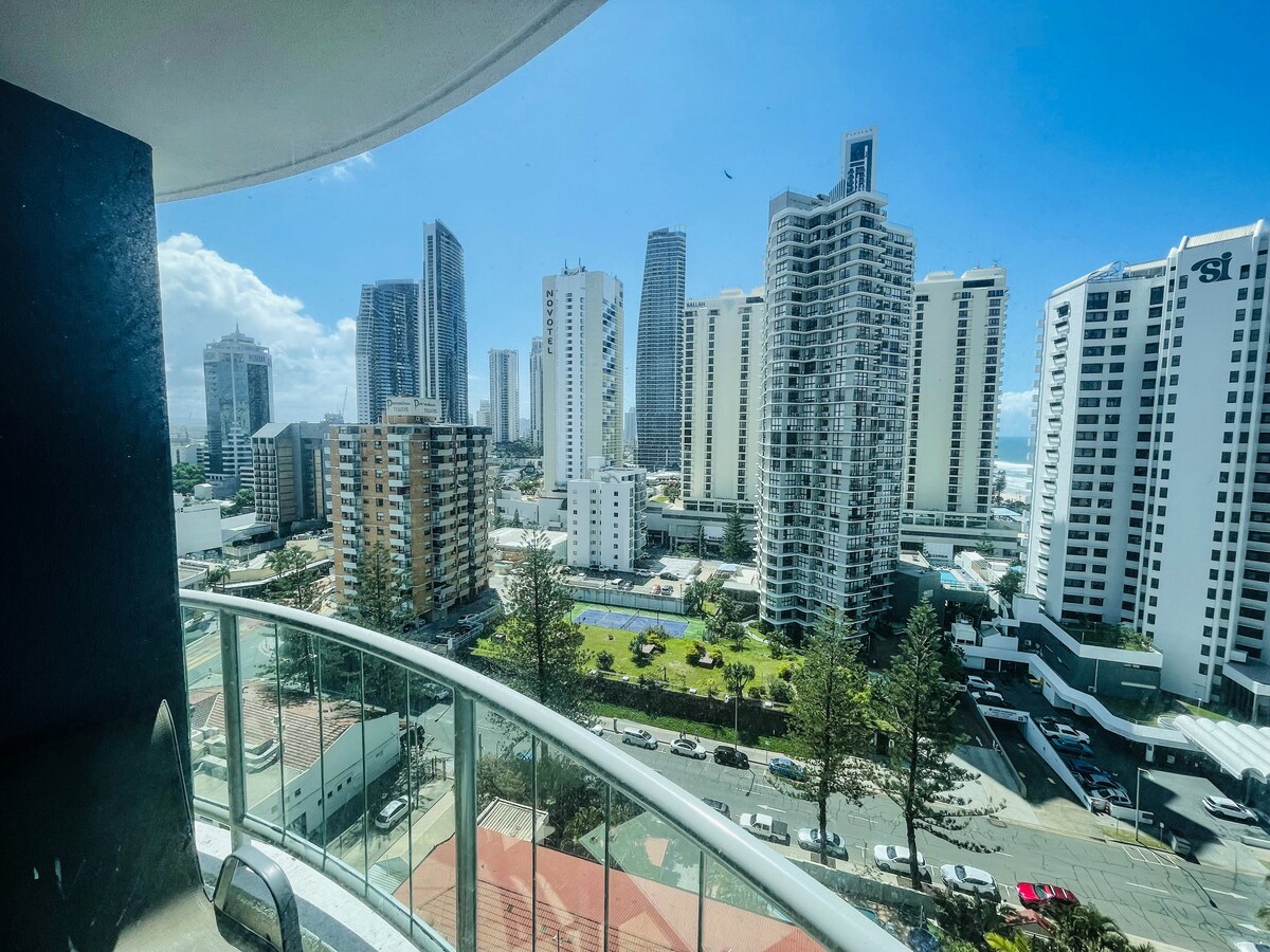 A panoramic view is captured from the balcony, showcasing the skyline of Surfers Paradise. Various high-rise buildings dominate the scene, while the vibrant greenery and outdoor pool can be seen below, offering a glimpse of the lively surroundings.