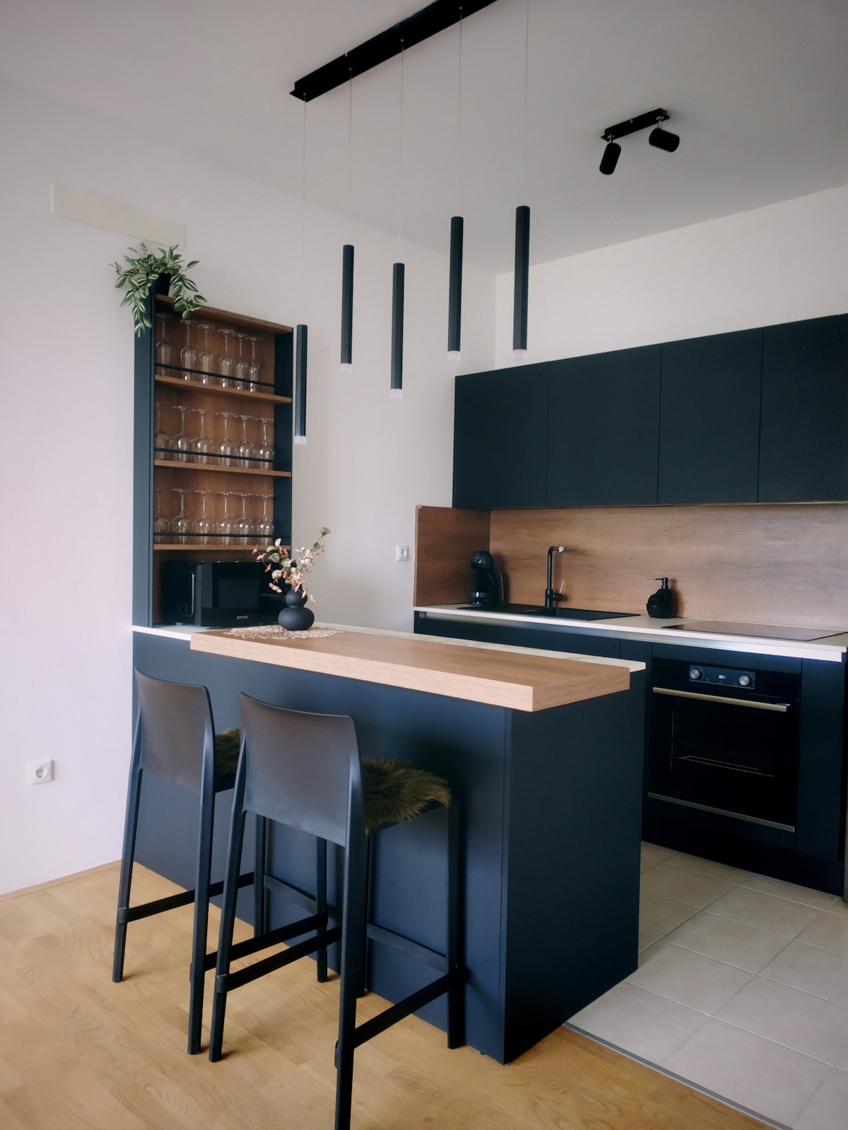 An open kitchen features dark cabinetry accented by a wood countertop. The bar area includes two high stools with a soft covering. Shelves display glassware, while modern appliances are positioned for convenience. Pendant lights hang from the ceiling, adding a contemporary touch.