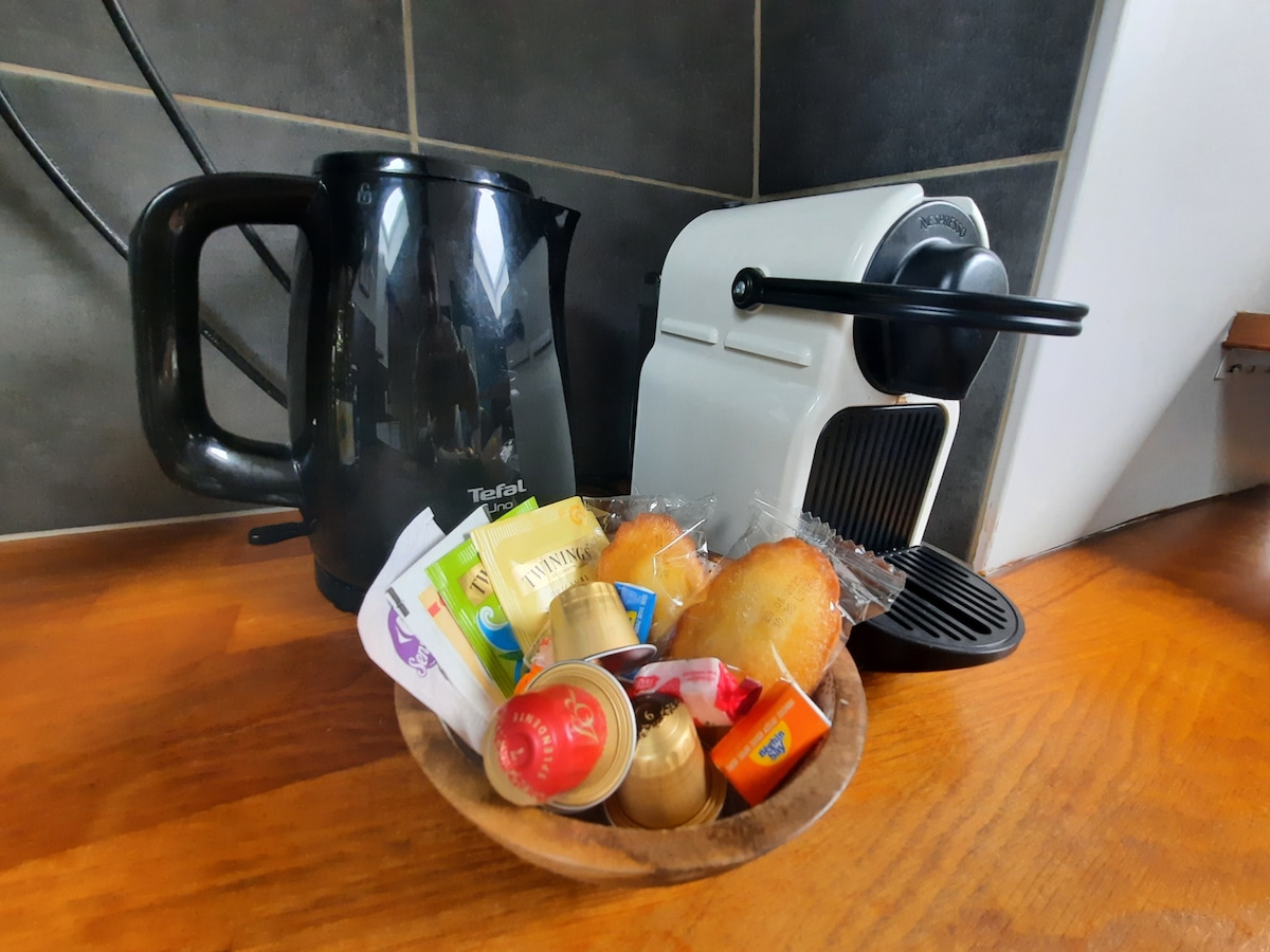 A kettle and a Nespresso coffee machine are positioned on a kitchen countertop. A wooden bowl holds assorted coffee capsules and sweet treats. Bright colors from the tea bags and coffee pods add visual interest to the setup.