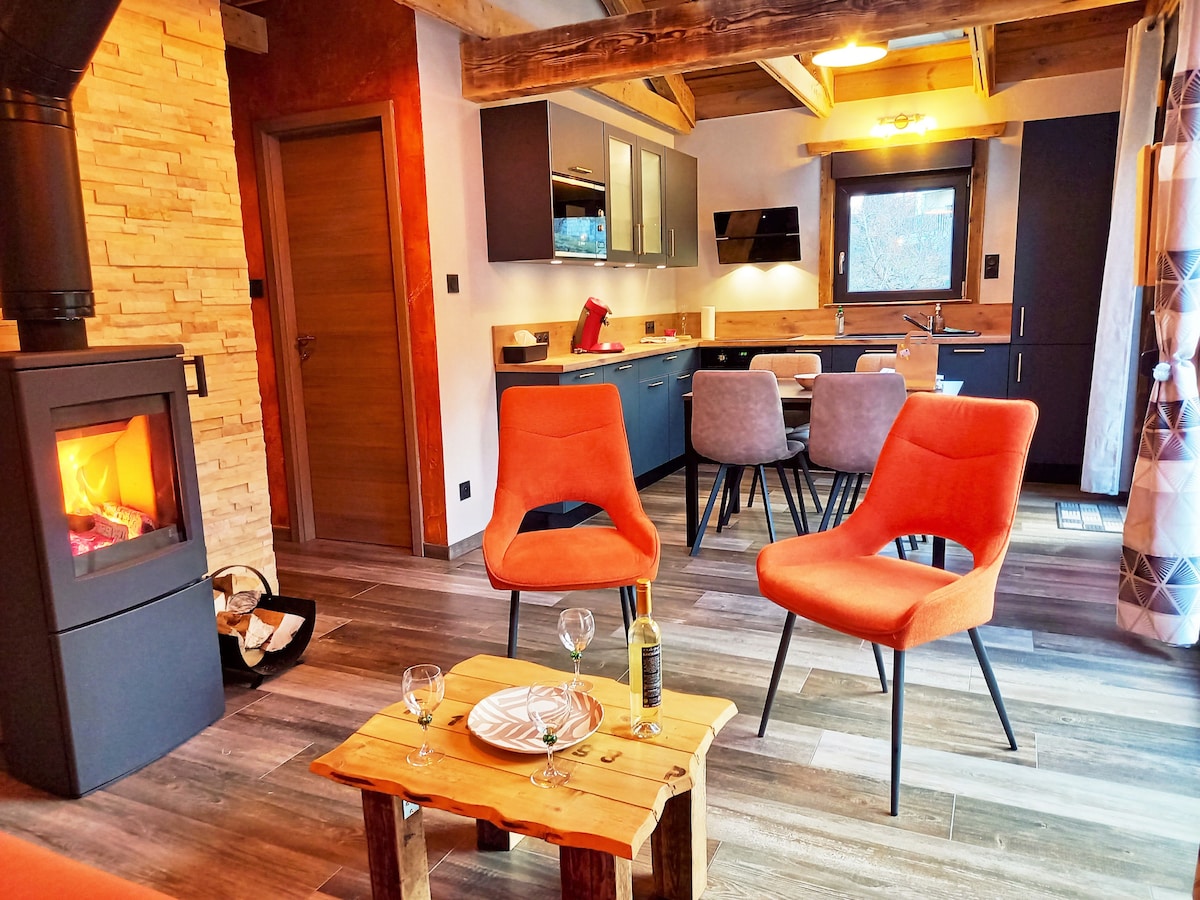 The living area features a wood stove surrounded by two orange chairs and a wooden coffee table. A modern kitchen can be seen in the background, with a dining table set for four. Light filters in through a window, enhancing the inviting space.