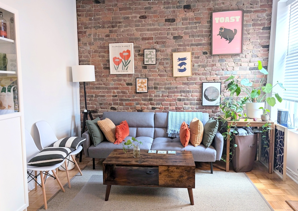 A cozy living area features a gray sofa adorned with cushions in earthy tones, set against a brick wall. Artwork, including a whimsical 'TOAST' print, decorates the wall. A wooden coffee table sits centrally, accompanied by a small green plant, and a bright window allows natural light to fill the space.