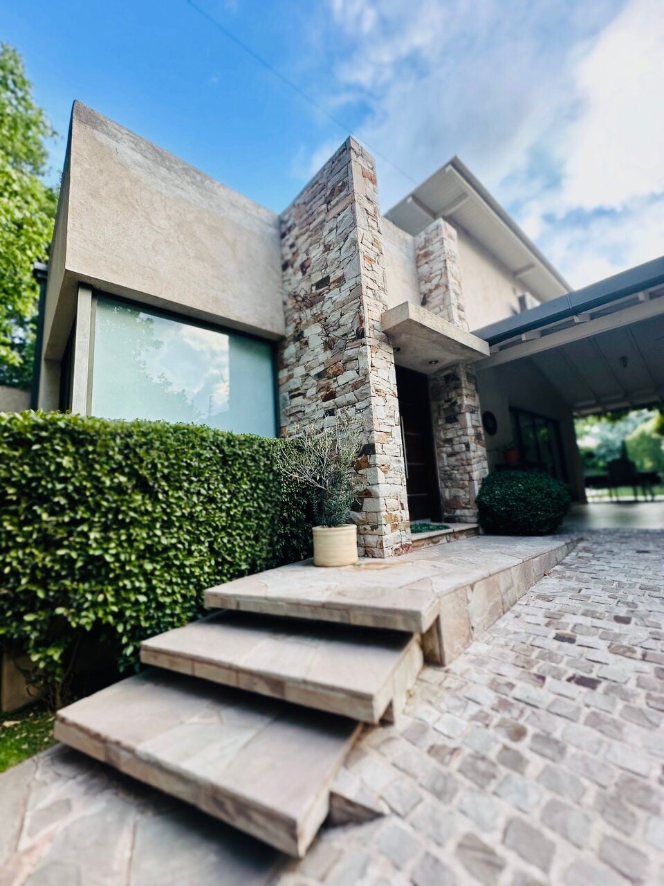 The entrance of the modern house is showcased, featuring well-crafted stone accents and large windows that allow natural light to filter through. Stone steps lead up to the front door, bordered by lush greenery that enhances the welcoming ambiance.