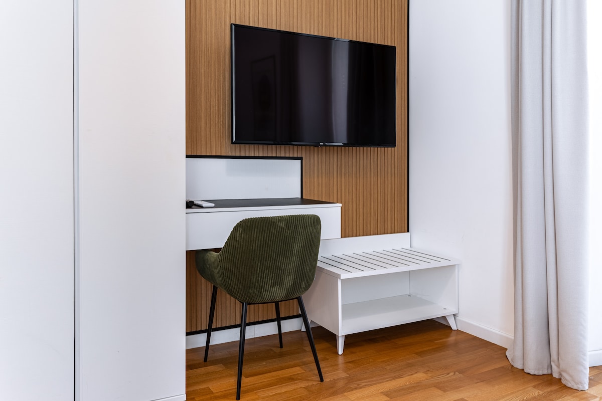 A modern workspace features a sleek desk and a comfortable green chair, positioned under a flat-screen television mounted on an accent wood-paneled wall. A white bench adds additional seating and storage below the television, complementing the clean lines of the room.