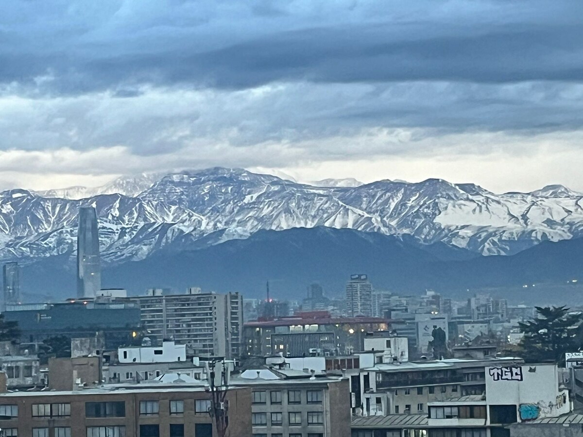 A stunning view showcases snow-capped mountains in the background, towering over the urban landscape. The city features a mix of modern buildings and rooftops, providing a contrast to the natural scenery, all under an expansive cloudy sky.