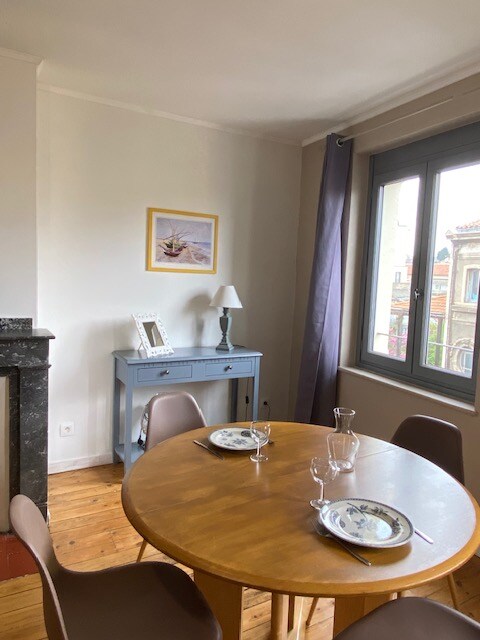 A dining area features a round wooden table set with plates and glasses. A small blue sideboard is positioned against the wall, complemented by a lamp and a framed picture. Natural light enters through a window dressed with a gray curtain, highlighting the warm wooden floor.