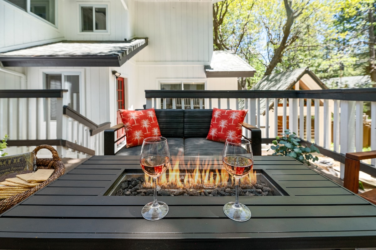 An outdoor seating area is shown, featuring a black sectional sofa adorned with decorative red throw pillows. A fire pit table with a flame sits in the center, complemented by two glasses of wine. Lush greenery and soft sunlight create a peaceful atmosphere.