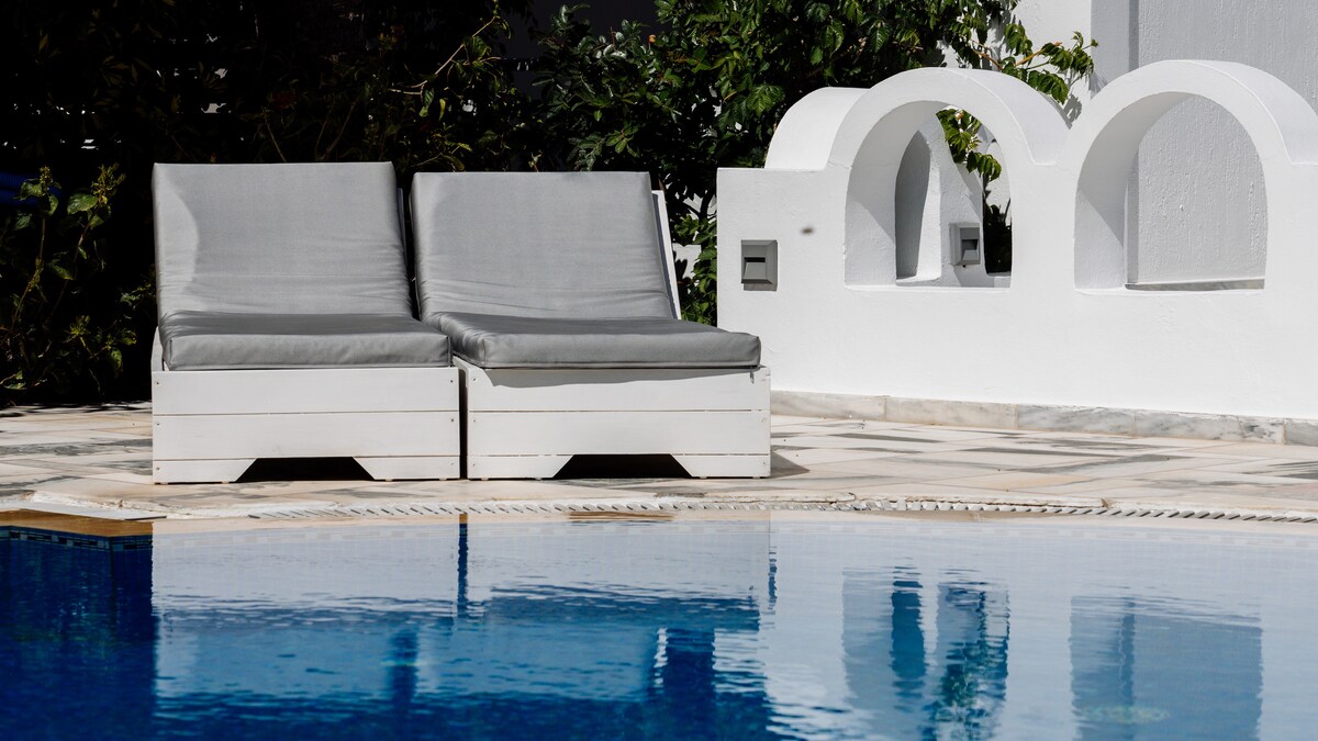 Two lounge chairs with gray cushions are positioned poolside, overlooking a clear blue swimming pool. Surrounding greenery provides a natural backdrop, while a white architectural feature complements the serene setting.