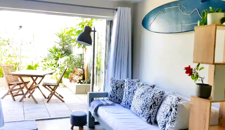 A light-filled lounge area features a spacious sofa adorned with patterned cushions, complemented by a small stool. A dining table with folding chairs and a vibrant garden can be seen through large glass doors, enhancing the connection to the outdoor space.
