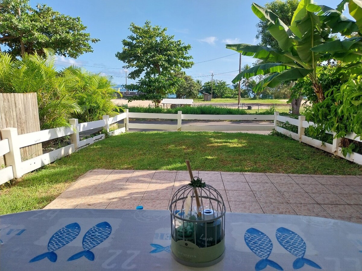 An outdoor view from the patio features a well-kept lawn with greenery and trees lining the property. A wooden fence frames the space, while a table with a decorative item sits in the foreground. The road is visible in the distance under a clear blue sky.