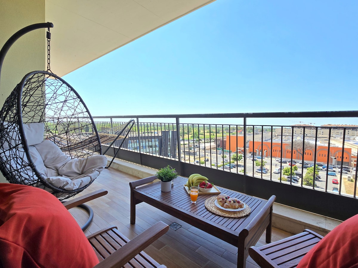An airy terrace is depicted, featuring a modern hanging chair and a wooden table set with refreshments. The view showcases a clear sky and a vibrant landscape, creating a space for relaxation and comfort. This area is designed for enjoying meals outdoors.
