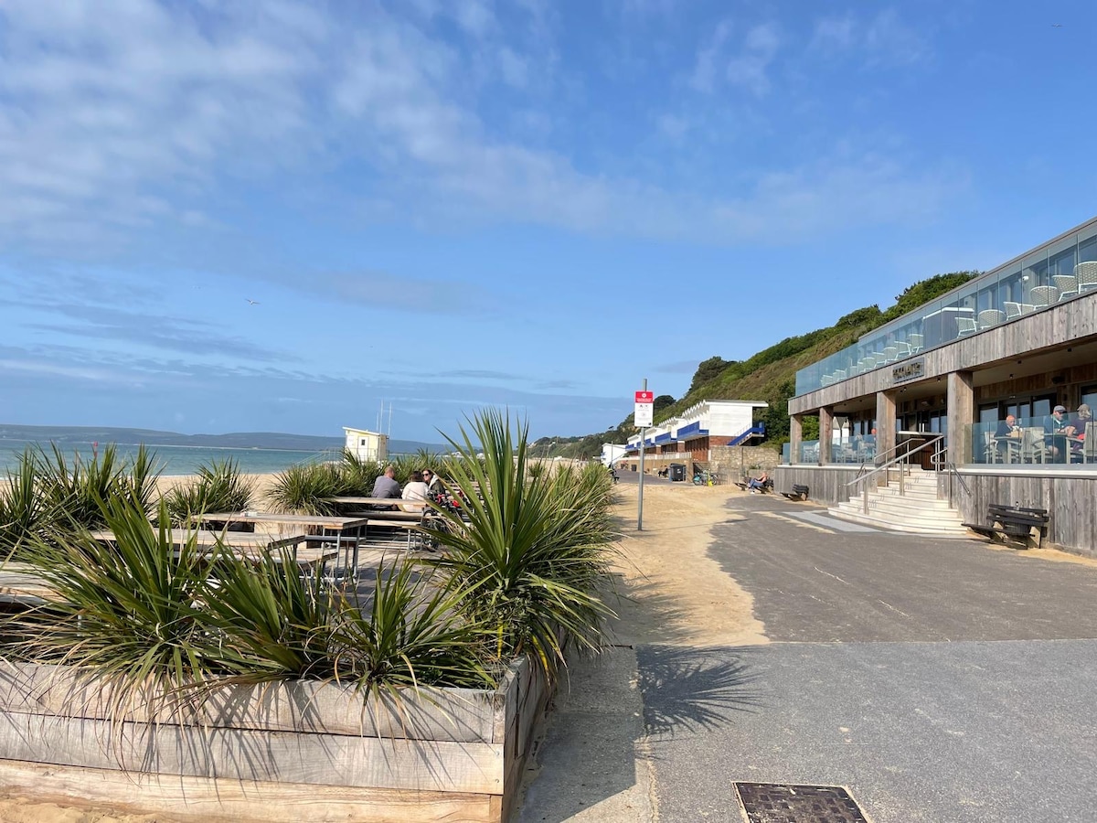 The image captures a seaside pathway lined with low wooden planters filled with green plants. A modern building with large glass windows is visible, which houses seating areas for outdoor dining. The ocean and a sandy beach stretch in the background under a partly cloudy sky.