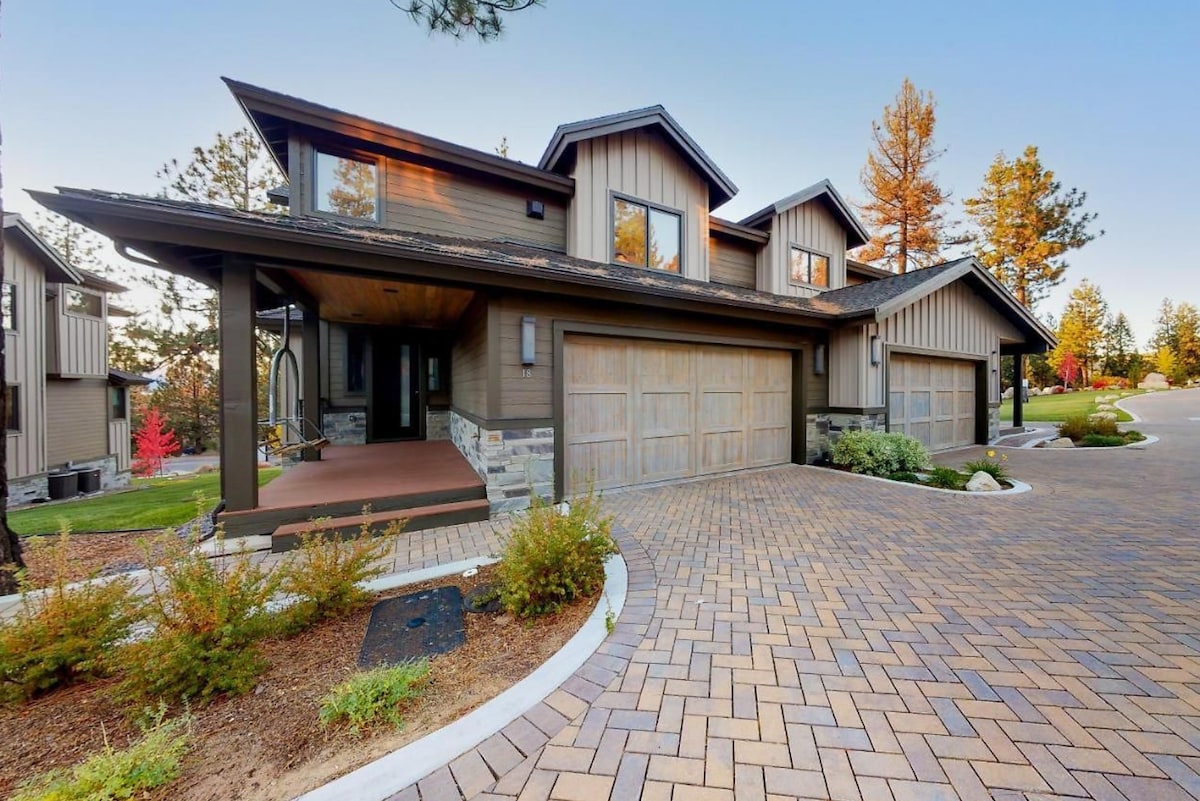 The exterior of the mountain villa is shown, featuring a two-story design with a broad front porch. A paved driveway leads to the attached garages. Landscaping includes various shrubs and trees, with an inviting atmosphere highlighted by the soft golden glow of the setting sun.