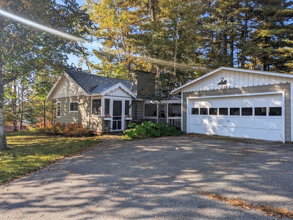 A charming cottage is seen surrounded by tall trees, featuring a light-colored exterior and a sizable garage with a white door. The gravel driveway provides ample space for parking, while sunlight filters through the leaves, creating a dappled light effect on the ground.