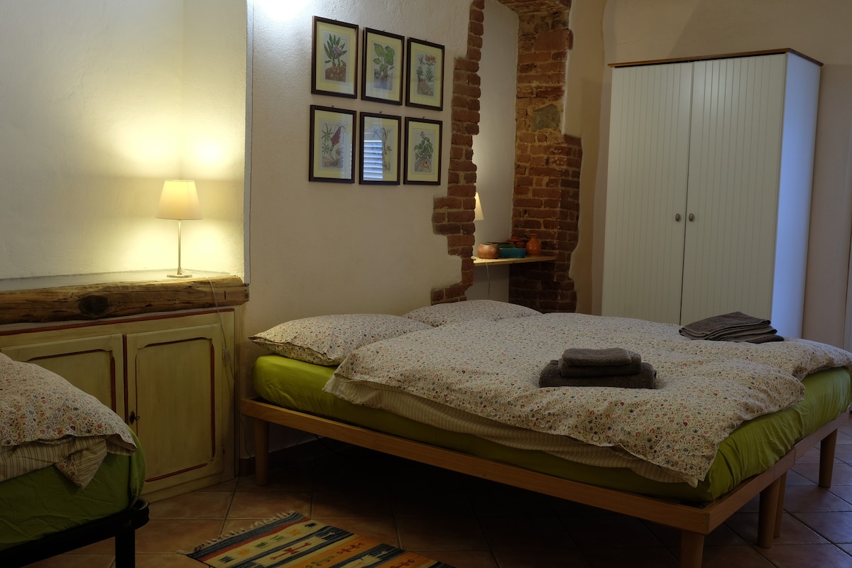 A cozy sleeping area is presented, featuring two spacious beds with floral-patterned bedding and neatly placed towels. A bedside lamp provides gentle illumination beside a rustic wooden cabinet. Artworks are displayed on the wall, and a white wardrobe is positioned in the corner.
