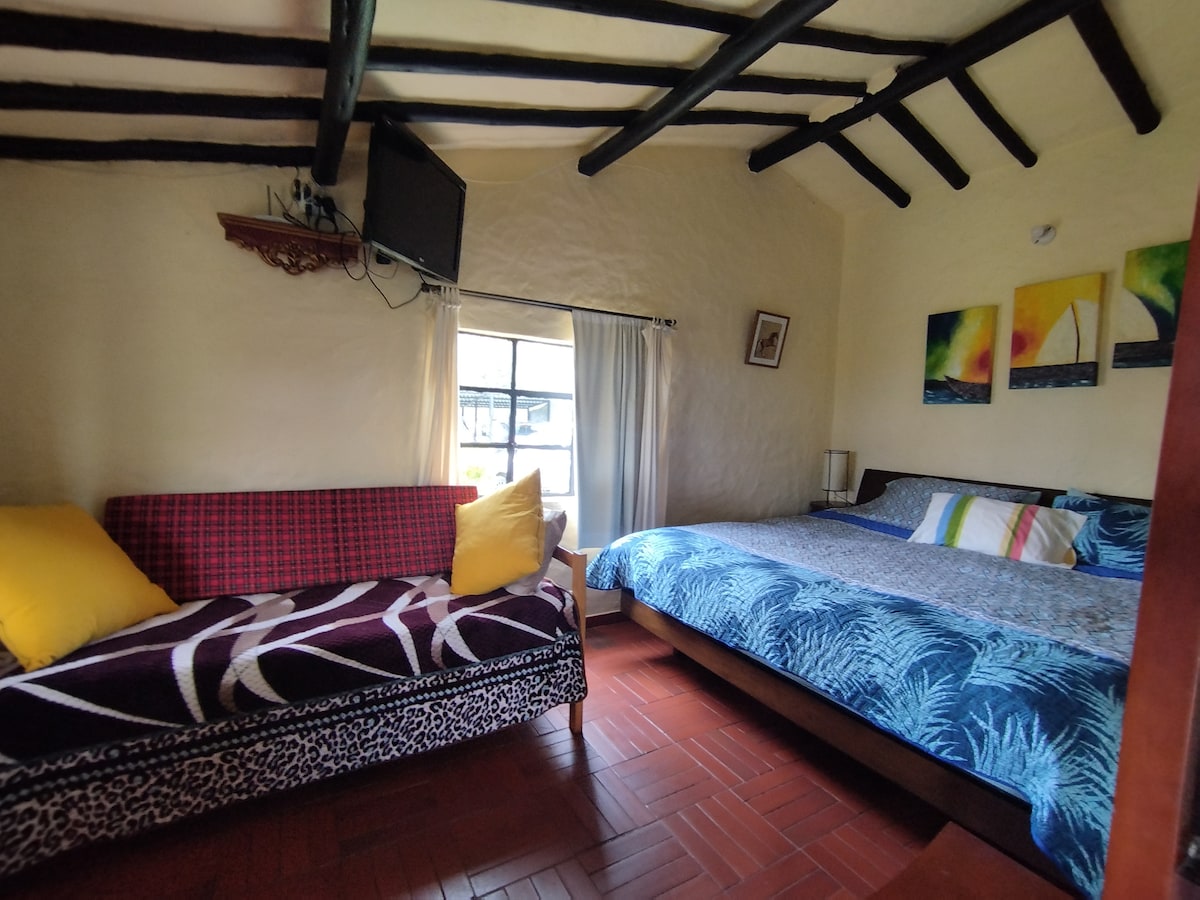 A sleeping area features a large bed with colorful bedding, complemented by a decorative wall art piece. A compact sofa bed with bright yellow cushions is situated nearby. Natural light enters through a window adorned with light curtains, illuminating the exposed wooden beams of the ceiling.