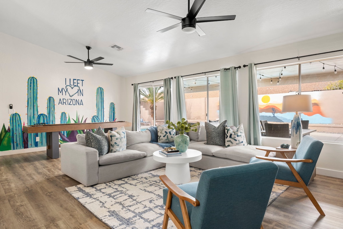 A bright, open living area features a large sectional sofa adorned with decorative pillows. A coffee table sits in the center, surrounded by two accent chairs. A mural of cacti decorates the wall, and large glass doors provide a view of the outdoor space.