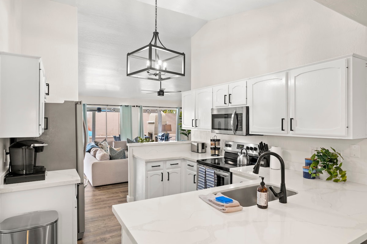 The kitchen features modern white cabinetry and sleek stainless steel appliances. A spacious countertop provides ample room for cooking, while a stylish light fixture hangs above. In the background, natural light illuminates the open layout connecting to a cozy living area.