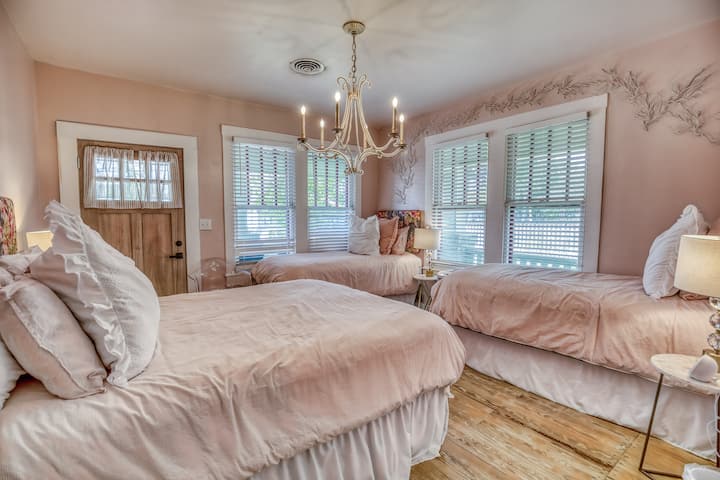 Spacious guest room featuring a queen bed and two twin beds, styled in a soft pink theme with a decorative chandelier overhead. A charming and whimsical space perfect for families or groups, blending comfort with a touch of elegance.