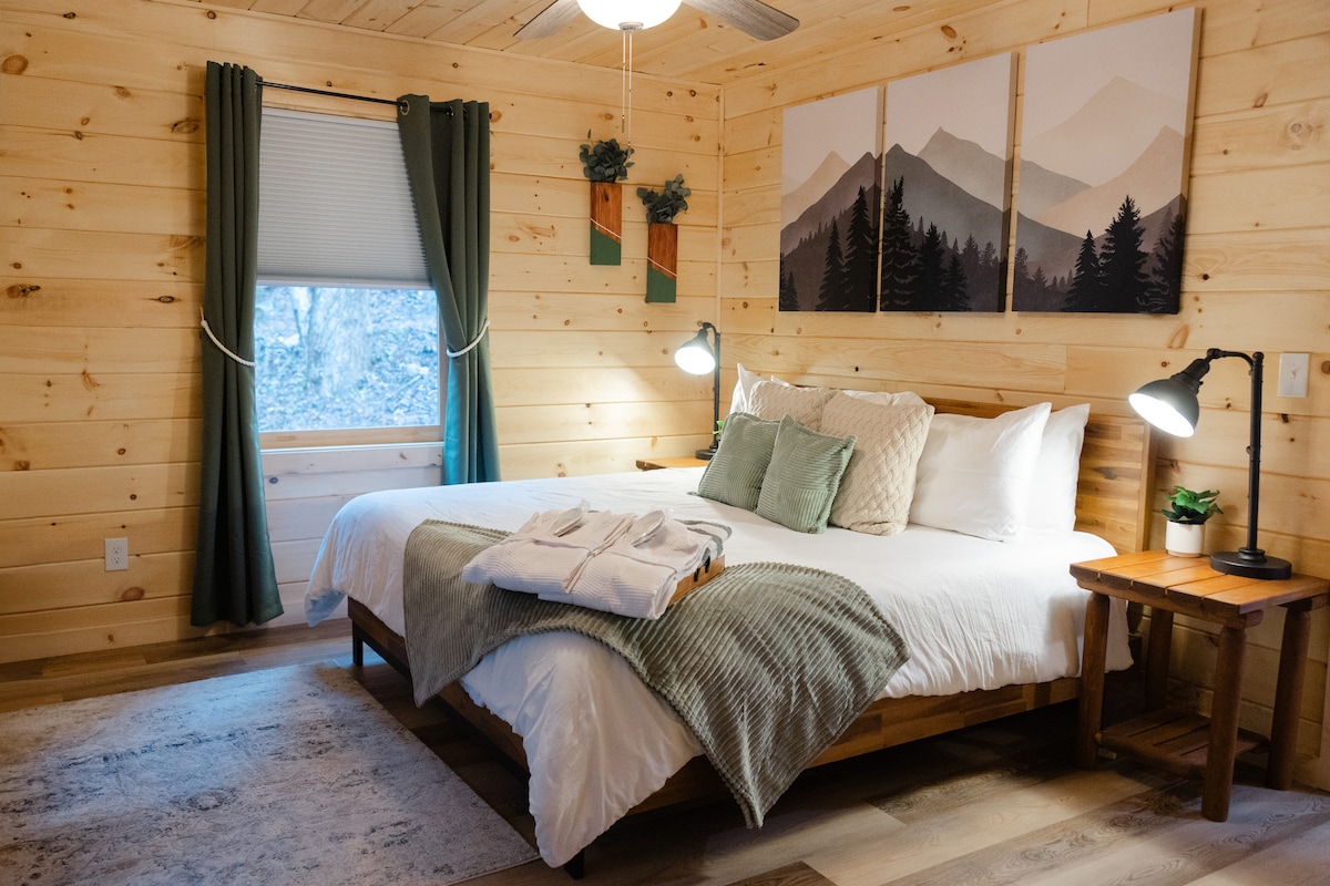 The bedroom features a spacious bed adorned with soft linens and decorative pillows, complemented by wooden bedside tables and reading lamps. Neutral-toned walls made of natural wood create a cozy ambiance, while a window framed by green curtains offers views of the surrounding nature.