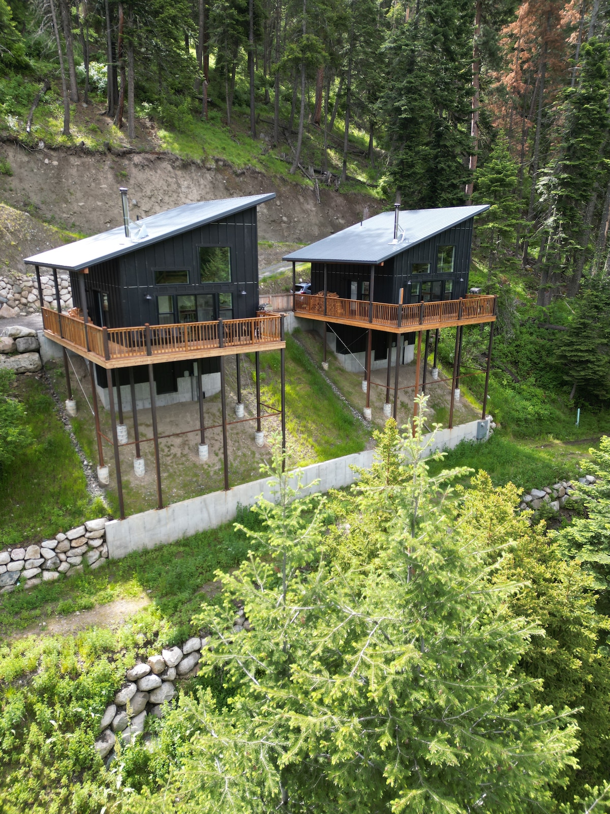 Two modern chalets are positioned on elevated platforms, surrounded by lush greenery. Each structure features large windows, allowing for ample natural light, and spacious semi-covered decks. The natural landscape and forest backdrop contribute to a serene mountain retreat setting.