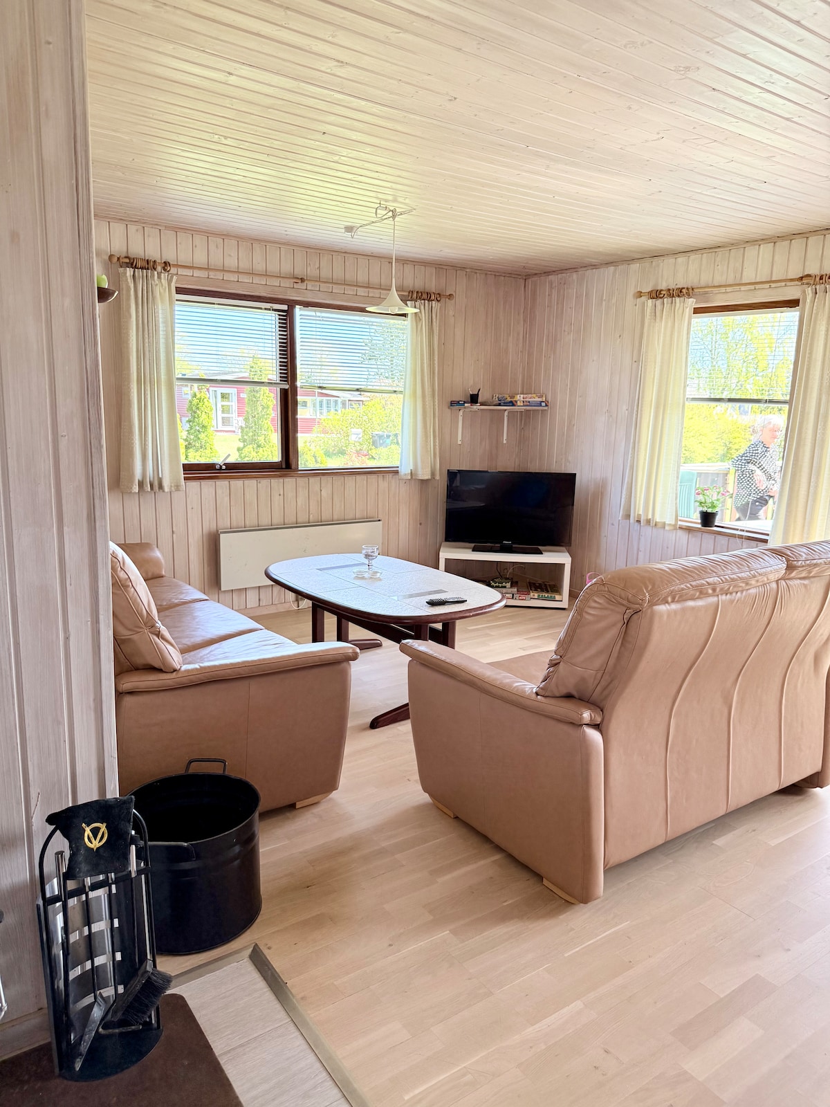 The cozy living area features two comfortable leather sofas facing a small circular table. A flat-screen TV is mounted on the wall, and natural light filters in through two windows, illuminating the wooden wall paneling and light-colored flooring.