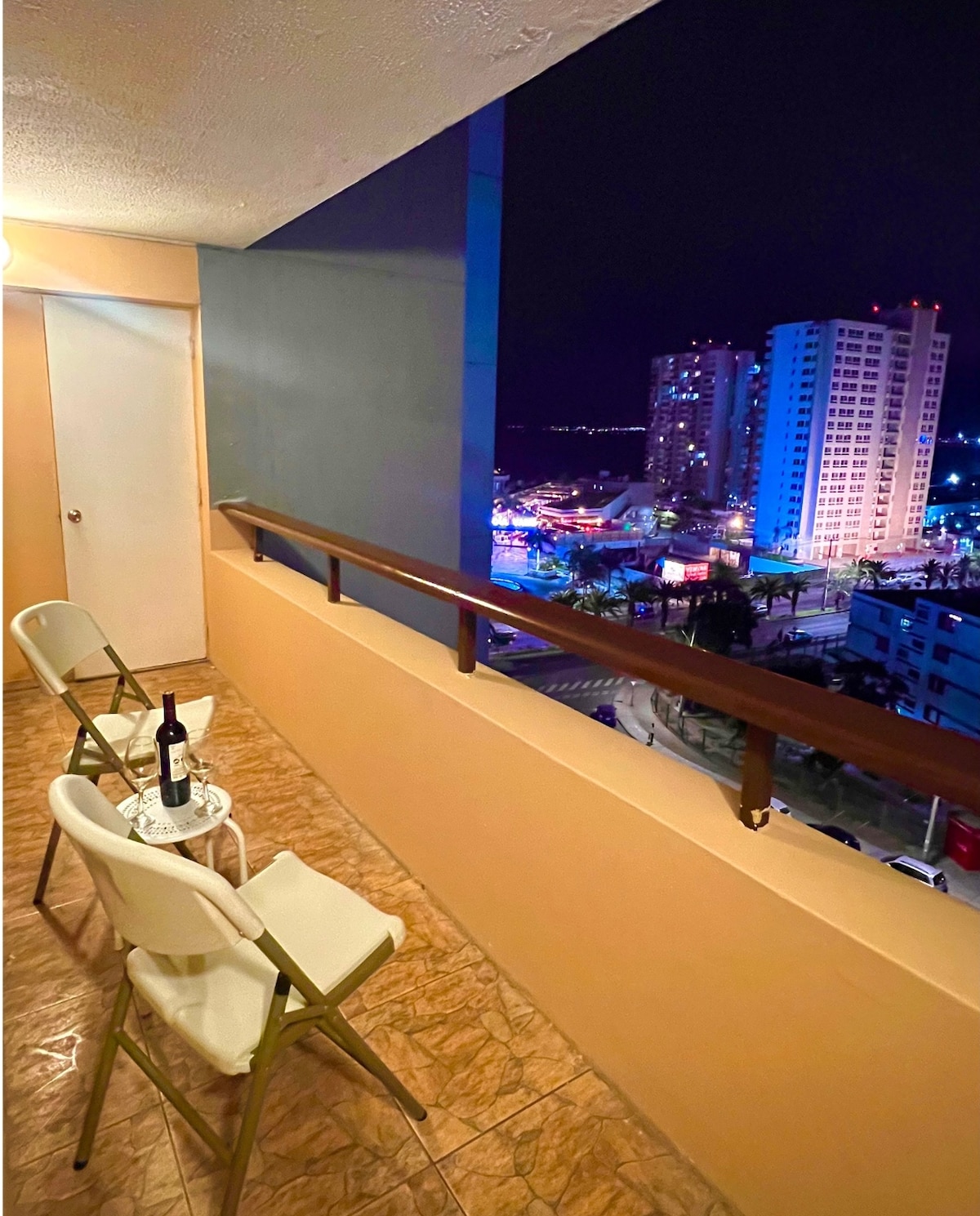 A balcony area is shown, featuring two folding chairs and a small table. A bottle of wine sits on the table. The view includes illuminated buildings and a vibrant cityscape, enhancing the atmosphere during the night.