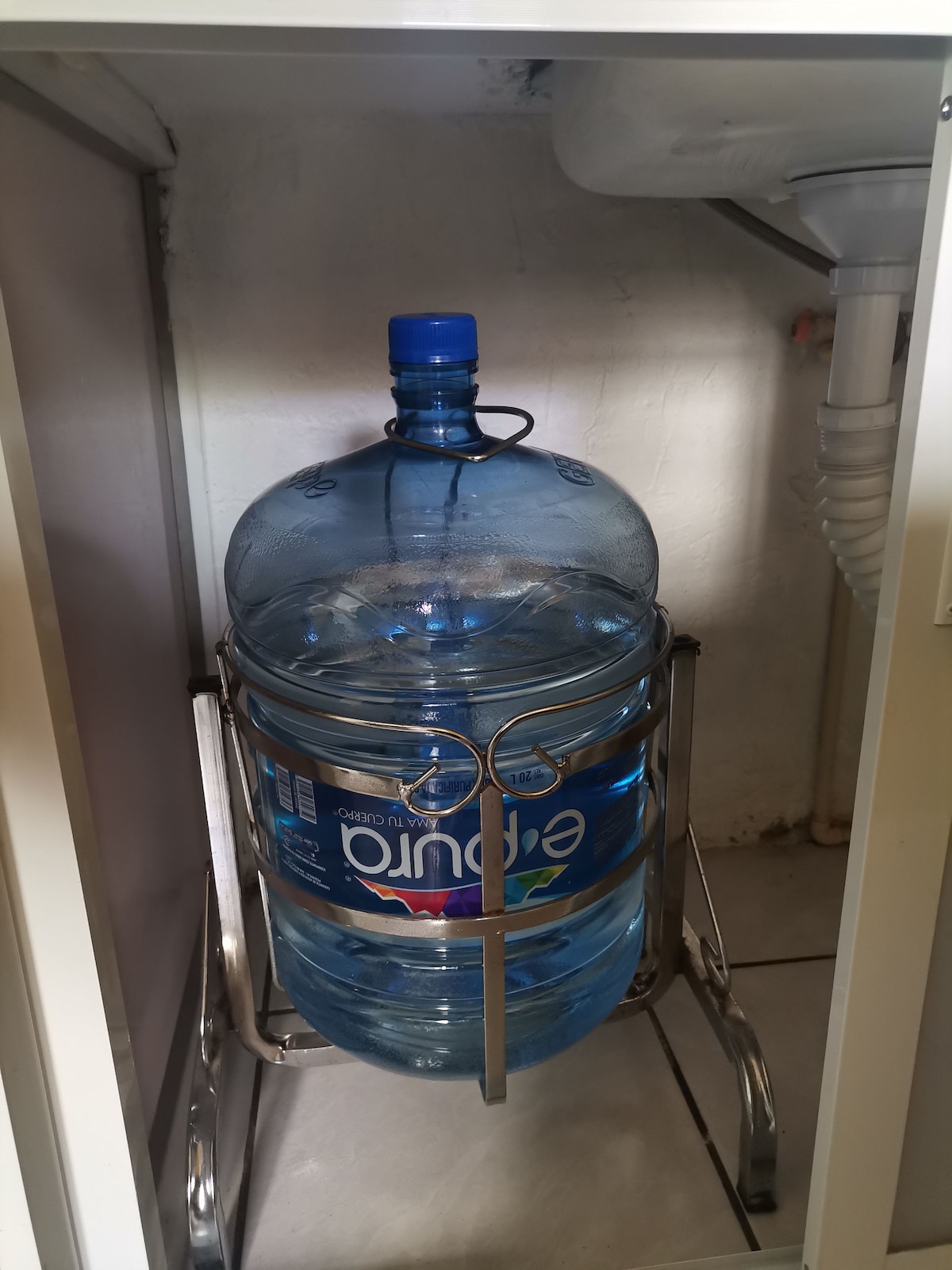 A large water bottle is securely placed in a metal stand beneath a sink. The bottle is clearly labeled and visible, providing access to refreshed drinking water.