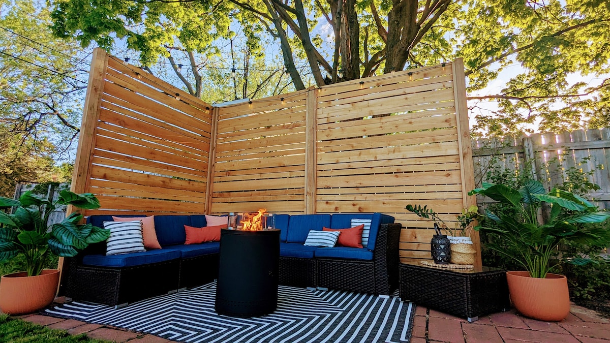 A cozy outdoor seating area is framed by wooden slats, providing privacy and comfort. A circular fire pit sits on a patterned rug, surrounded by plush seating adorned with colorful cushions. Potted plants add a touch of greenery, enhancing the inviting space.