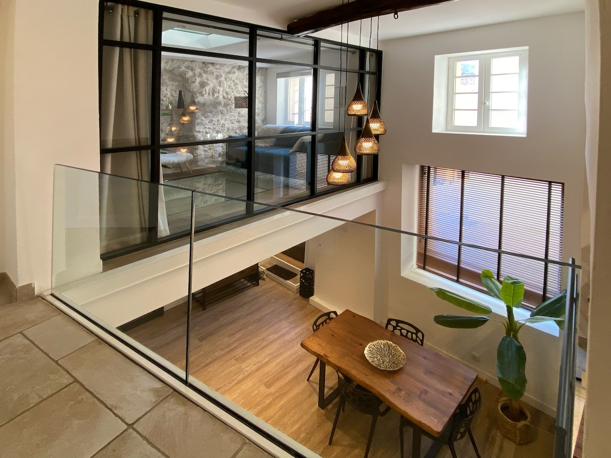 A modern living space with a glass railing overlooks a cozy dining area with a wooden table and decorative bowl. Pendant lights hang from the ceiling above, while natural light filters through large windows, illuminating the stone wall and contemporary furnishings in the background.
