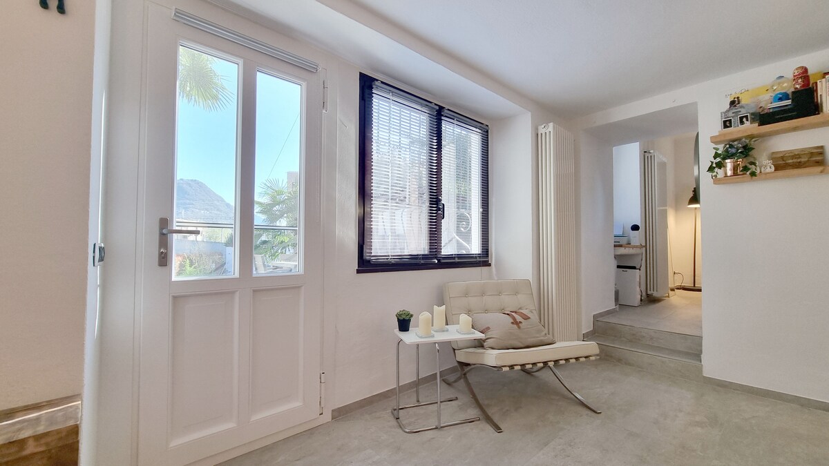 A bright, open living area features a modern chair positioned by a window, providing natural light and a view of greenery outside. The walls are painted in a light hue, and simple decorative elements are visible on shelves in the background, enhancing the serene environment.