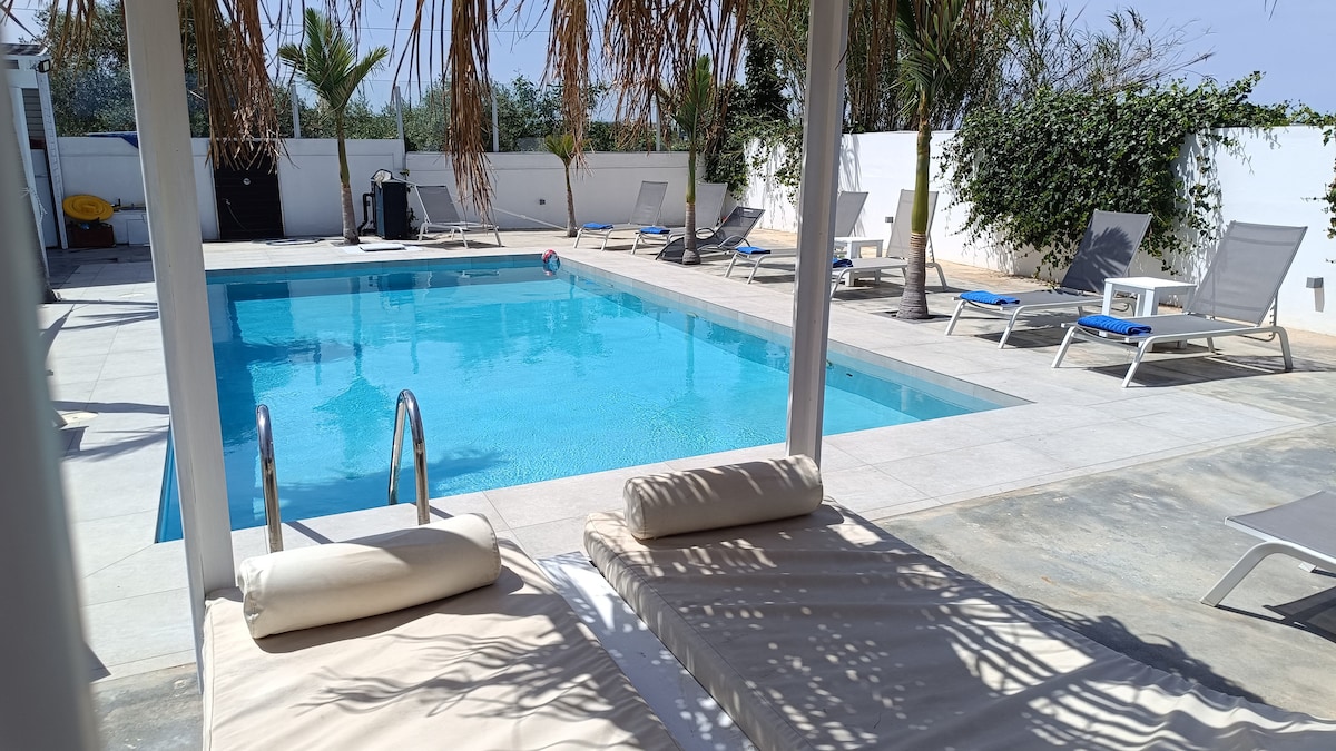 A private pool area features a spacious swimming pool surrounded by sun loungers. Palm trees provide shade, and a covered seating area is visible nearby. The surroundings are enclosed by a wall, ensuring privacy for guests enjoying the outdoor space.