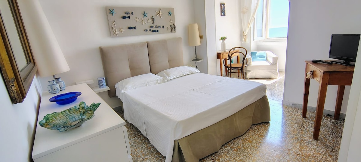 חדר שינה כפול גדול עם ריהוט מודרני וכמה פריטי עתיקה - ברקע, אזור שולחן וחלון המשקיף על הים. 