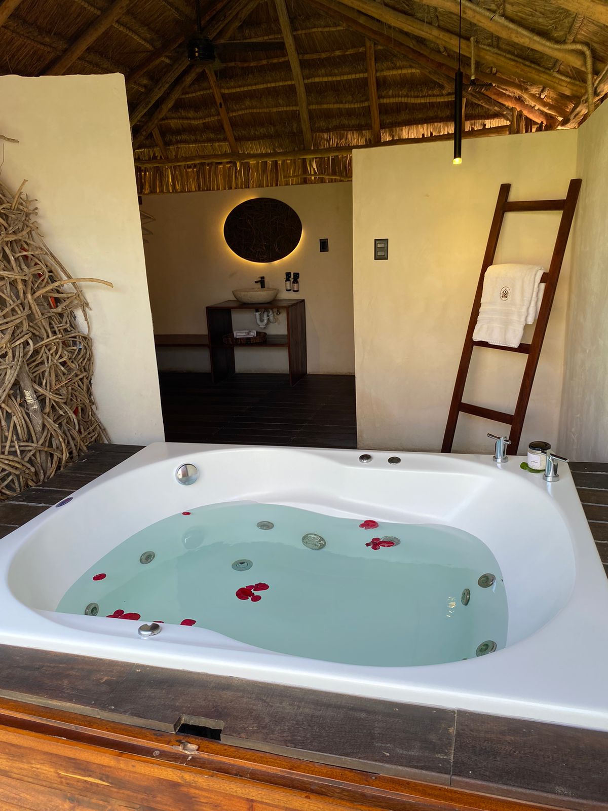 A private tub is situated indoors, set against a rustic backdrop. The tub contains clear water adorned with rose petals and stones. A wooden ladder leans against the wall, and a modern sink area is visible in the background, framed by natural materials.